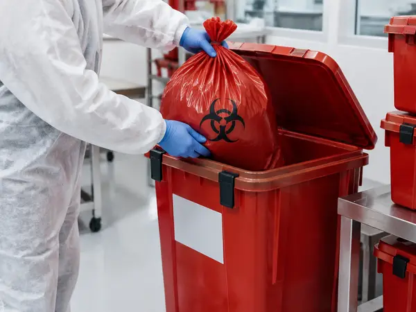 Gloved technician placing a sealed red biohazard bag into a rigid lidded waste container